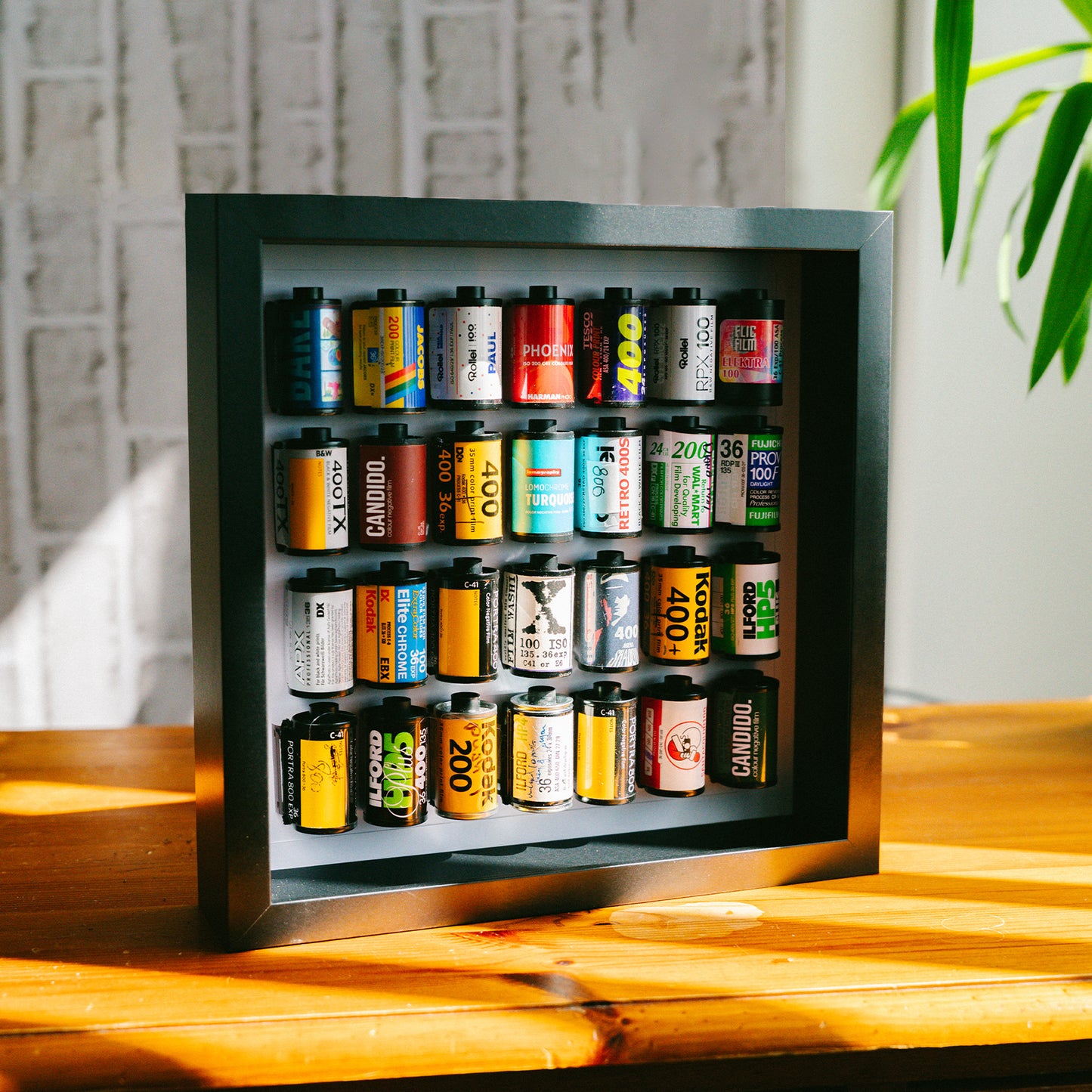 Retrozi signature film reel wall art — a black shadow box frame displaying a curated grid of 20 colourful 35mm film canisters including Kodak Gold 200, Kodak 400, Fujifilm, Candido and Phoenix, photographed on a warm wooden surface with natural light. Unique personalised film photography gift and home decor.