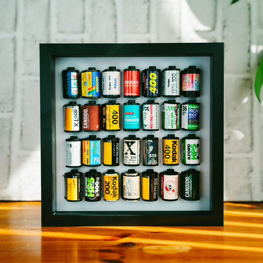 Collection of 35mm film canisters displayed in a framed shadow box, featuring brands including Kodak, Fujifilm, Ilford, Cinestill, and Candido, arranged in four rows on a warm wooden surface.